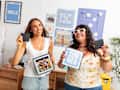 Two women smiling and holding photo frames in a colorful, decorated room with beach-themed wall art.