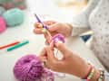 A close-up view of a woman’s hands as she skillfully crochets with purple yarn. The background includes colorful crochet hooks, balls of wool, and a pincushion, showcasing an organized and vibrant crafting environment. The bright workspace highlights her focus and creativity.