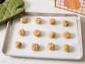 Cookie dough balls with chocolate chips arranged on a baking sheet lined with parchment paper, green oven mitt nearby.