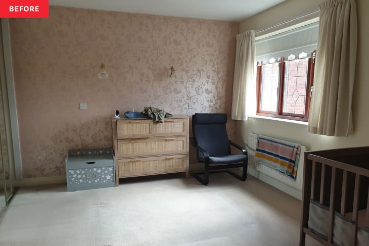 Spacious room with floral wallpaper, a wooden dresser, a gray storage box, a black chair, and a crib by the window.